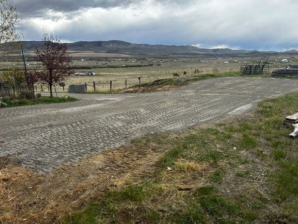 Gravel road leading to open fields and mountains under a cloudy sky. Green grass on the sides.