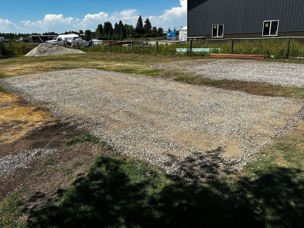 Gravel lot preparation for construction with grassy edges and nearby buildings.