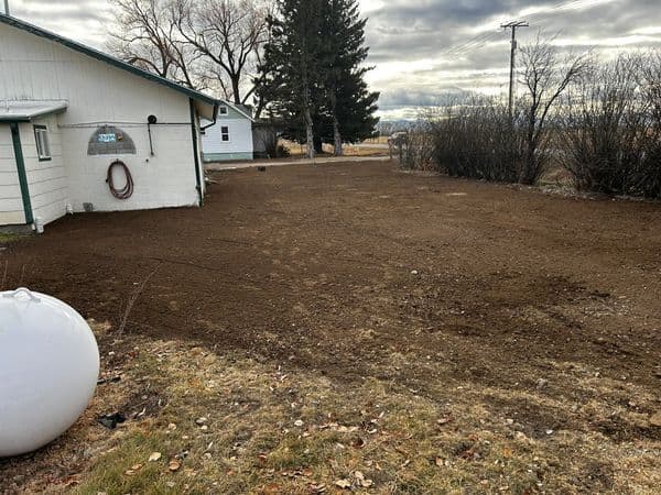 Cleared yard space with freshly tilled soil, surrounded by trees and a house.