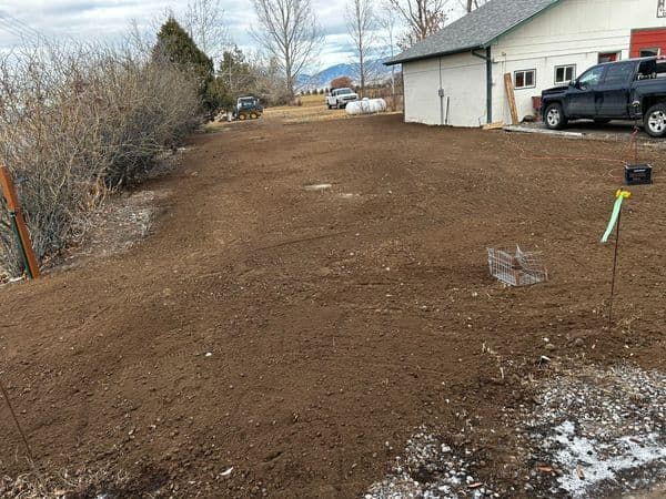 Bare dirt area ready for landscaping near a garage, surrounded by trees and vehicles.