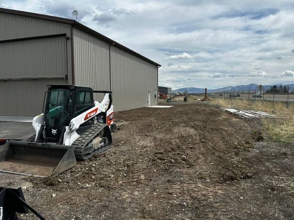 Bobcat skid steer on cleared land beside a gray industrial building under a cloudy sky.