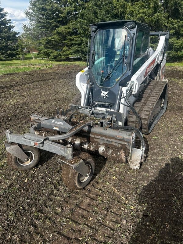 Bobcat skid steer loader with soil tiller attachment on freshly tilled land.