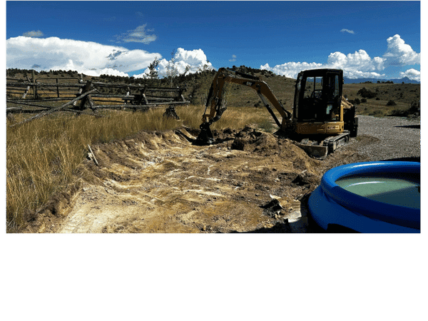 Excavator working on a dirt path near a pool in a rural landscape under a blue sky.